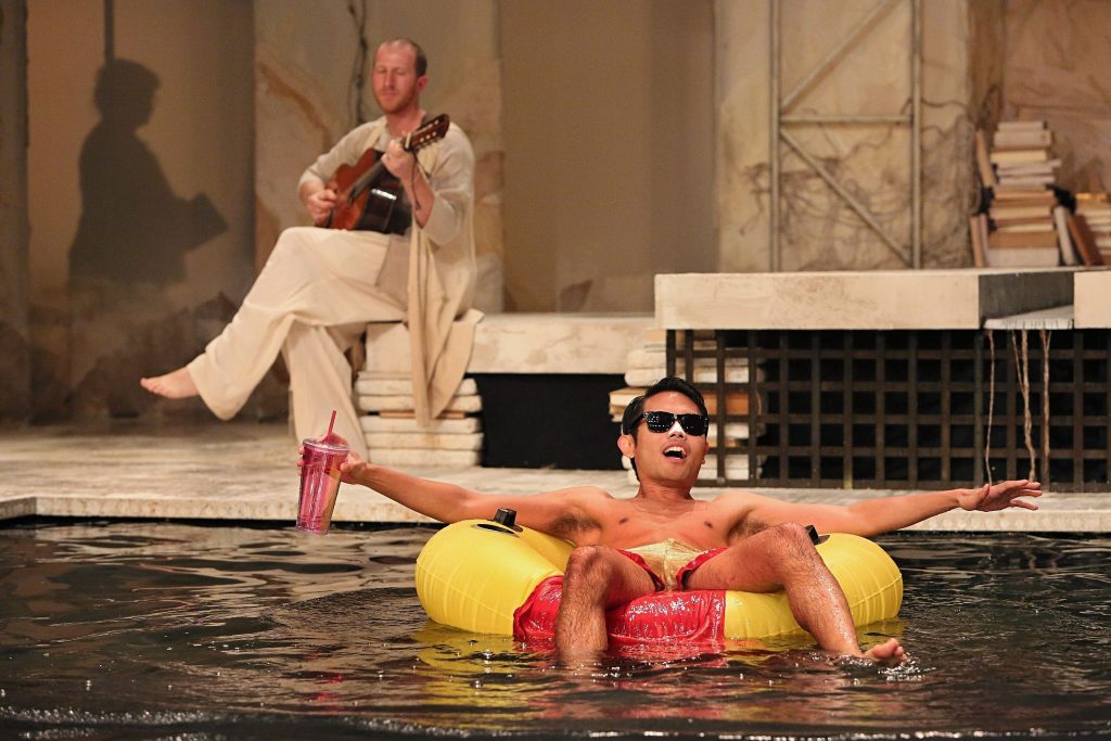 Ari Picker and Nathaniel Claridad in "Metamorphoses." Picker is further back on the stage with a guitar, while Claridad is floating on a tube in a pool of water as part of the stage.