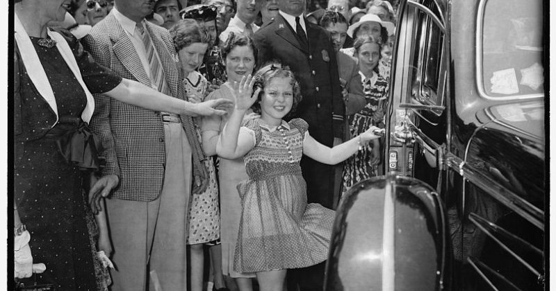 A young Shirley Temple waves at the camera as she gets into a car