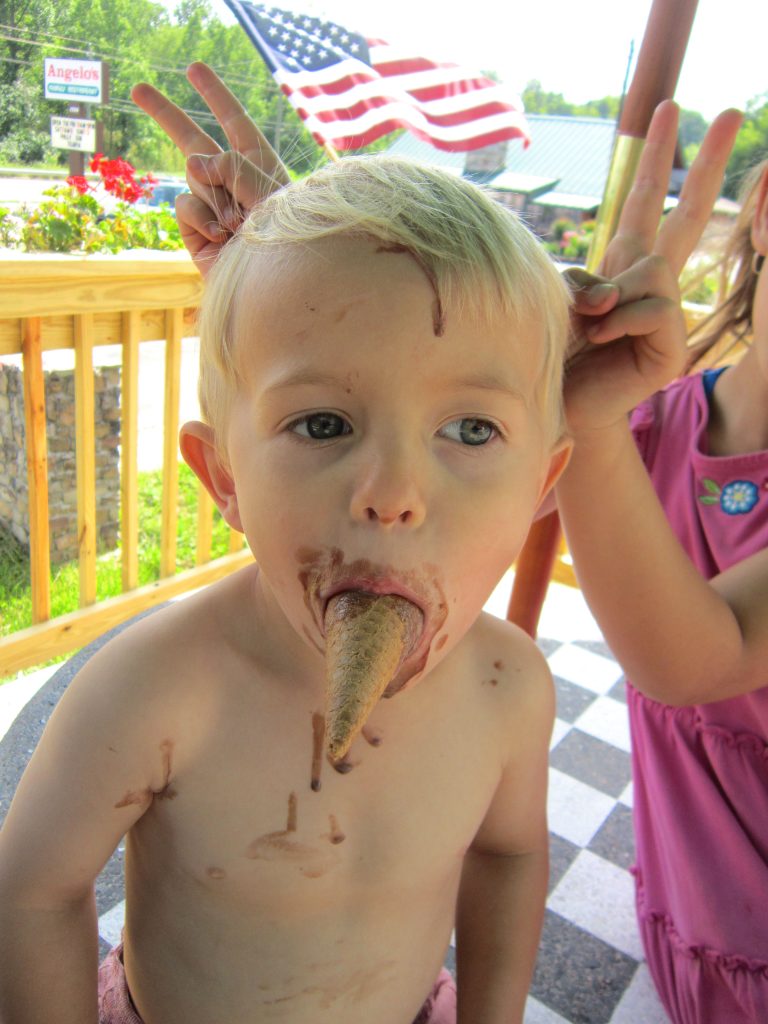 A young boy eats ice cream -- with the cone sticking out of his mouth. A young girl (not seen) gives him bunny ears on his head