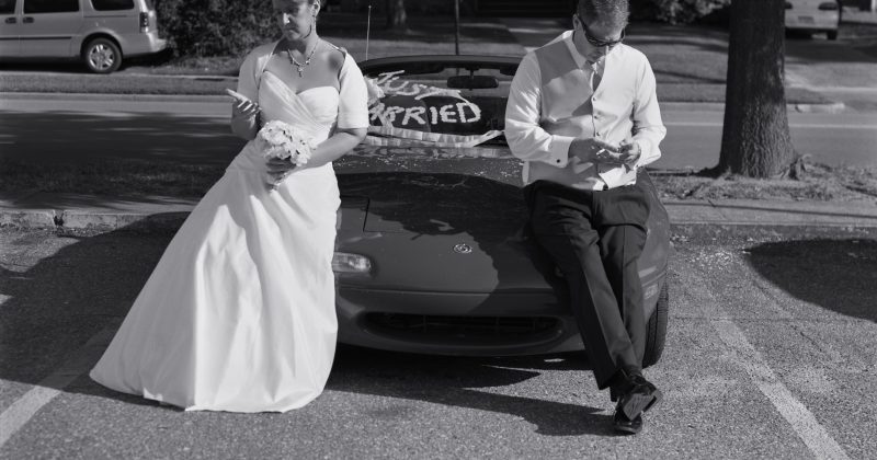 Black and white photo of a newlywed couple sitting on a car, posed as though looking at their phones -- though the phones are removed