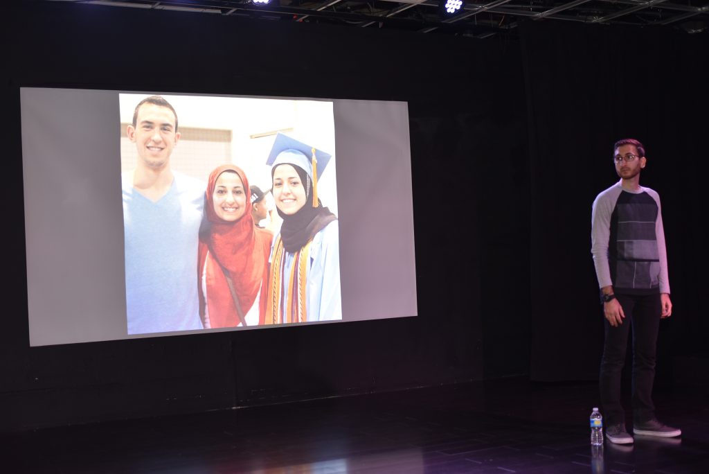 Mohammad Moussa in front of a screen featuring an image of Deah Barakat, Yusor Abu-Salha, and Razan Abu-Salha