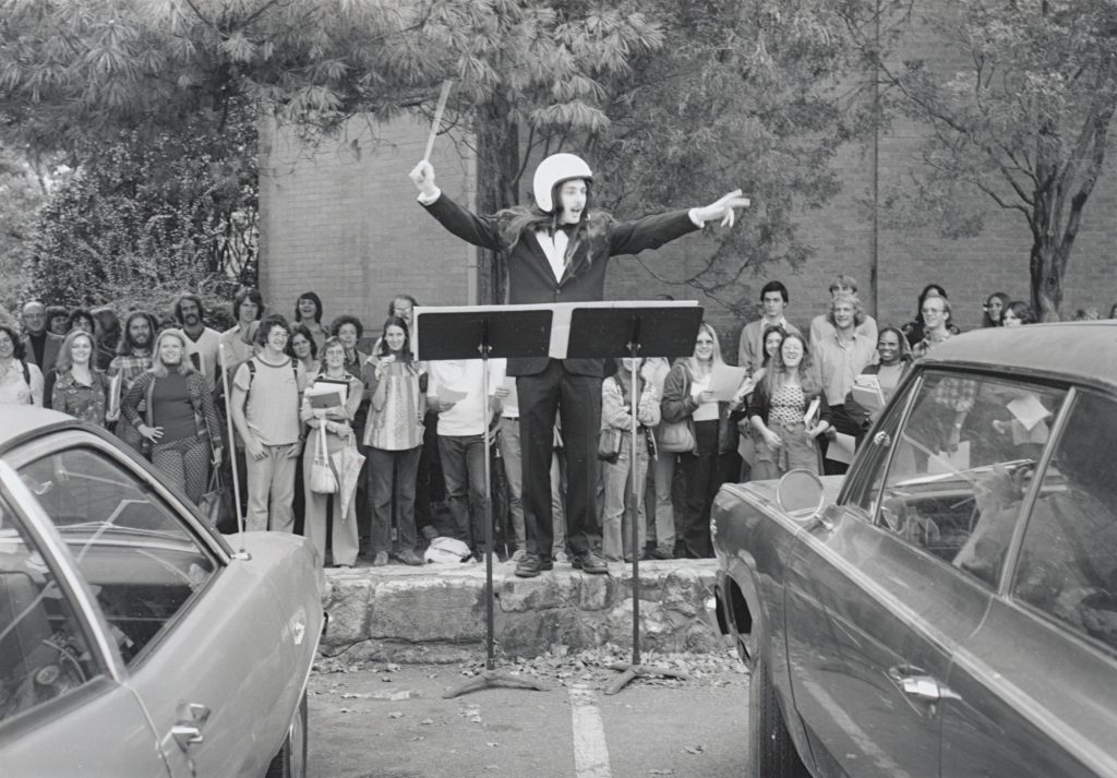 A student, wearing a helmet, stands on a short stone wall and conducts music to cars. A crowd of people stands behind him