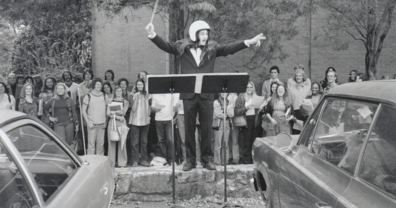 A student, wearing a helmet, stands on a short stone wall and conducts music to cars. A crowd of people stands behind him