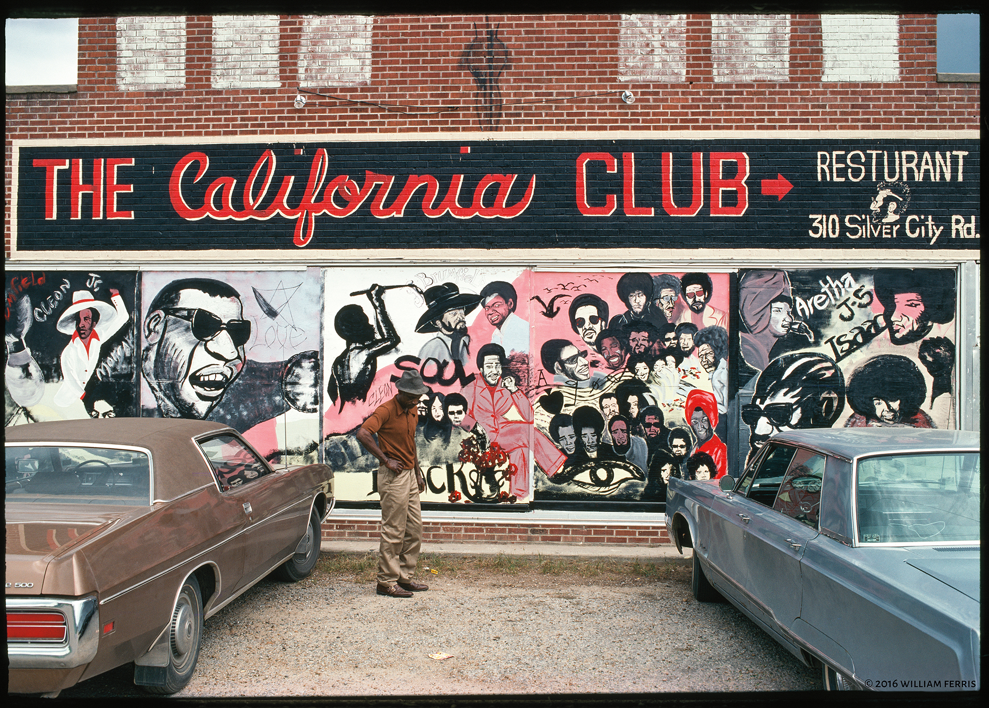 A man stands between two cars outside the California Club, which is decorated with a mural