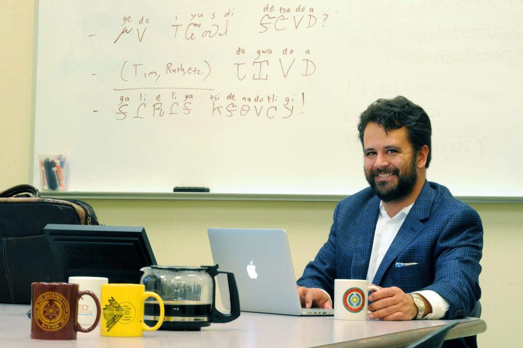 American studies assistant professor Ben Frey at his computer with a coffee pot and surrounding mugs. Words in Cherokee is written on the board behind him.
