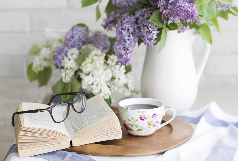 Image shows a wooden plate with an open book on it and a pair of glasses sitting on it. Beside the book is a flowered tea cup. A white vase pitcher sits behind the plate with purple and white flowers in it.