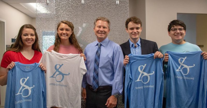 College Dean Kevin Guskiewicz stands in the center of a group of four students, all holding Synergy Unleashed T-shirts.