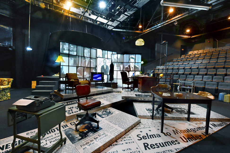 The Edges of Time set in the Paul Green Theatre. The floor is decorated with large images of historic newspapers, including those from the Civil Rights Movement. Three desks are set up in different corners, as well as a chair near a small television. The back display has multiple screens, which show historic clips of various news anchors.