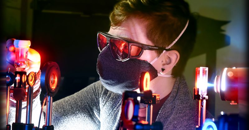 Rachel Bangle works in the laser lab with a mask on and pulsing lights around her.