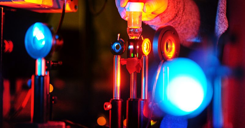 Rachel Bangle adjusts a device in the laser labs in Murray Hall. Bright blue lights reflect, against a warm orange glow.