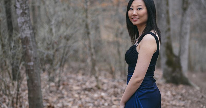 Clara Yang pictured smiling in a dress in the woods, surrounded by trees and leafs on the ground