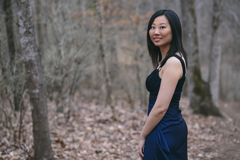 Clara Yang pictured smiling in a dress in the woods, surrounded by trees and leafs on the ground