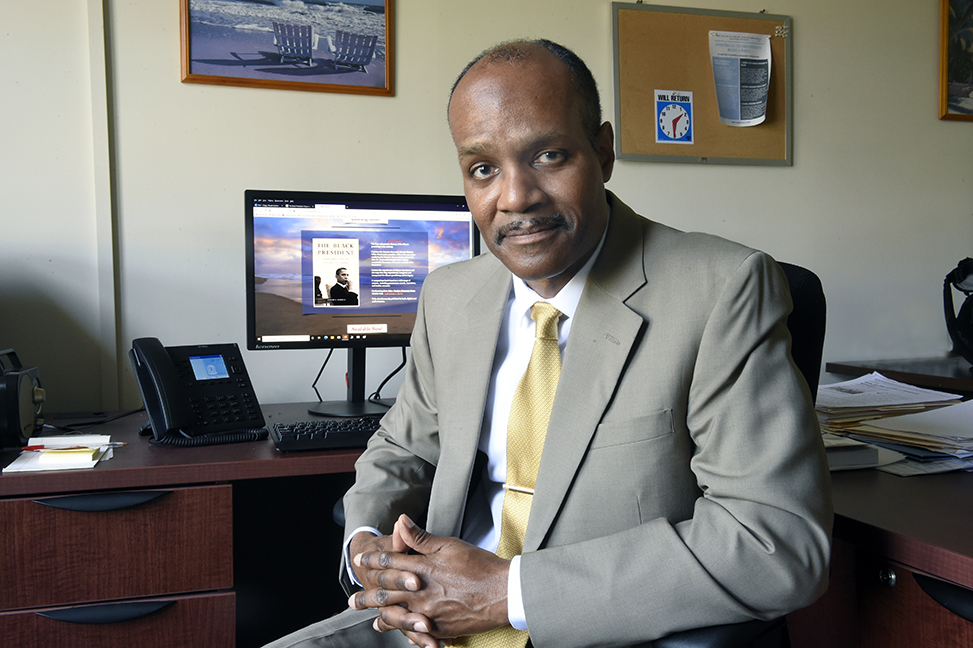 Claude Clegg, dressed in a suit, sits at his desk and smiles at the camera with a computer screen in the background with a picture of his Obama book cover.