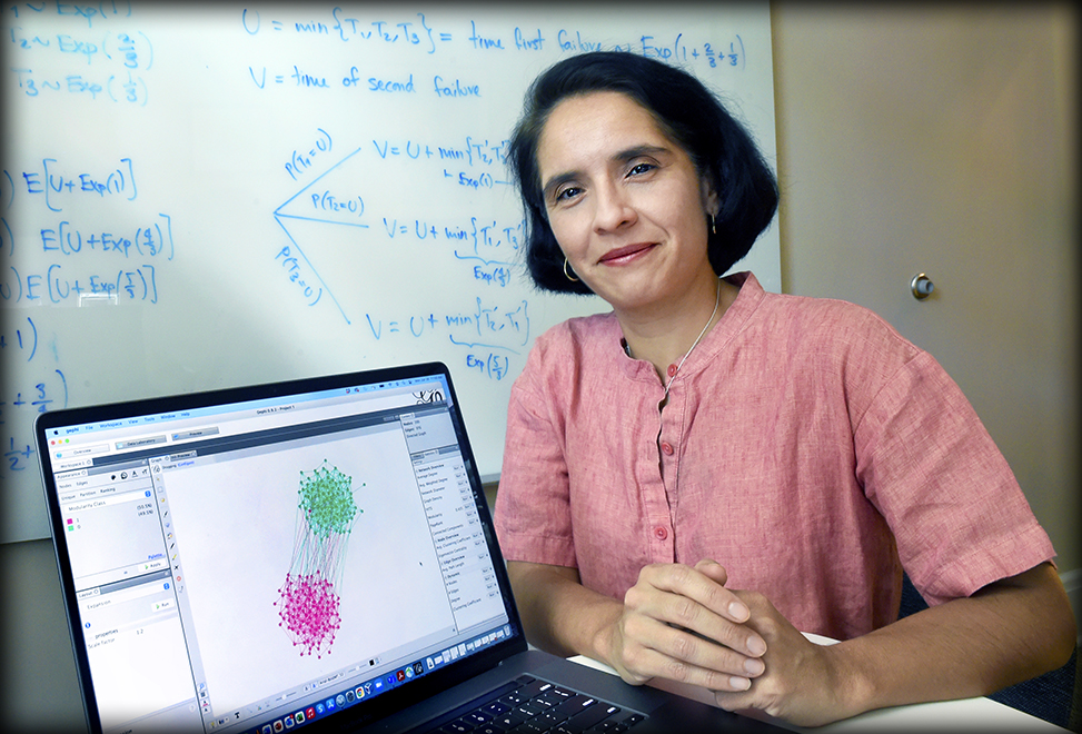 Mariana Olvera pictured smiling beside her computer showing a mathematical model. She is sitting in front of a whiteboard filled with formulas.