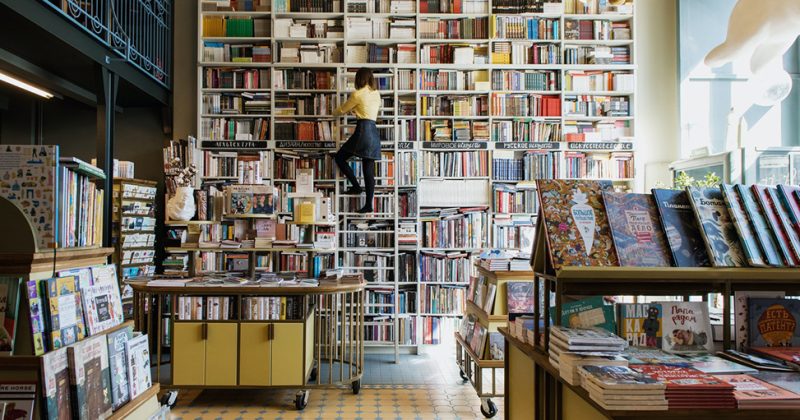 A women on a ladder in a bookstore surrounded by shelves of books.