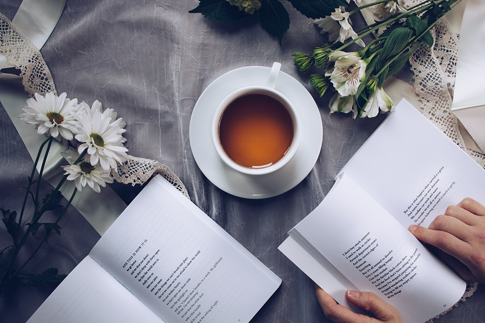 Two open books and a hand pointing to writing in one book. A cup of coffee sits on the table with the books along with some flowers.