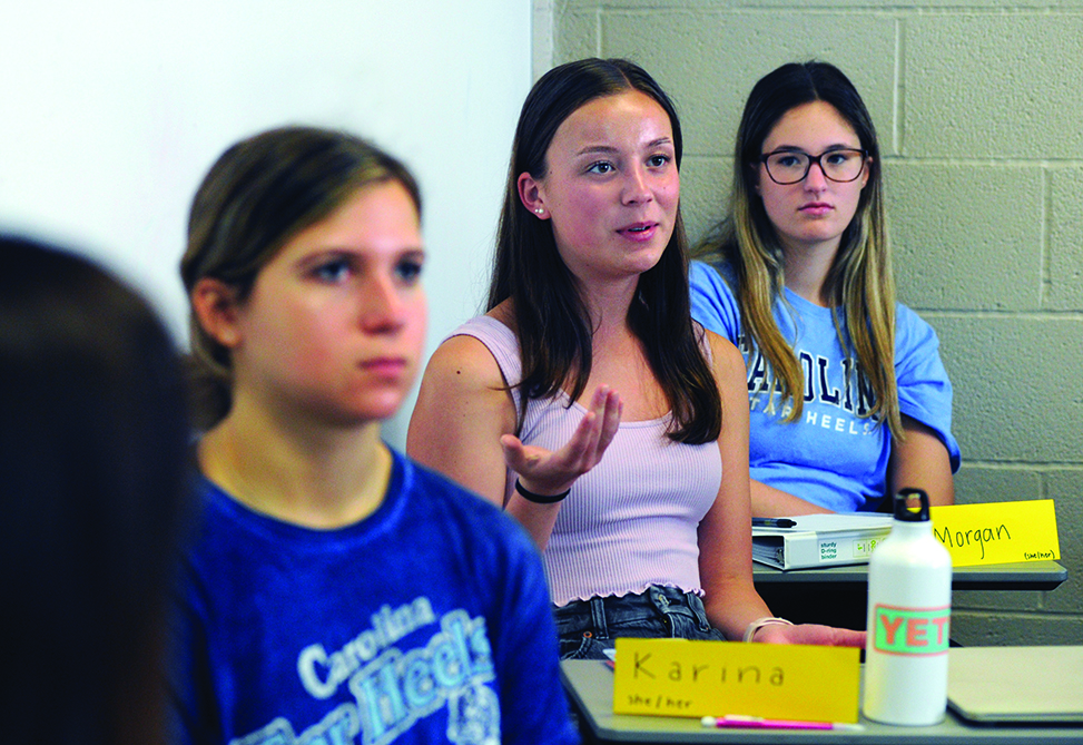 Three students sit at their desks, one gesturing while speaking.
