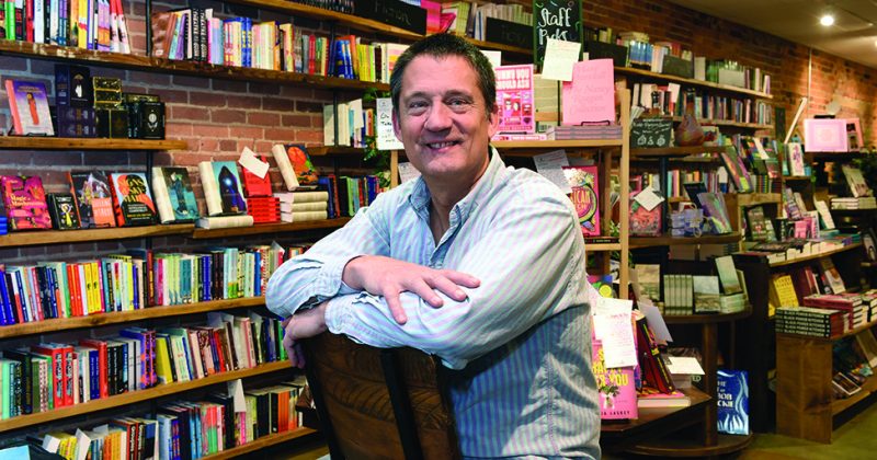 Fitzhugh Brundage sits backward on a chair, leaning on the backrest, in Epilogue Books. Shelves and displays of books fill the background.