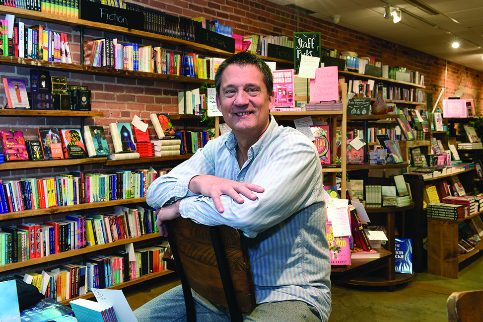 Fitzhugh Brundage sits backward on a chair, leaning on the backrest, in Epilogue Books. Shelves and displays of books fill the background.