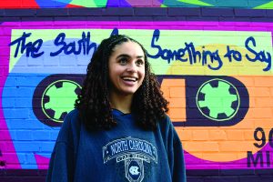 Hallie Brew wears a Carolina sweatshirt and looks to her left. Behind her is a colorful mural featuring a cassette tape and the words, "The South's Got Something to Say."