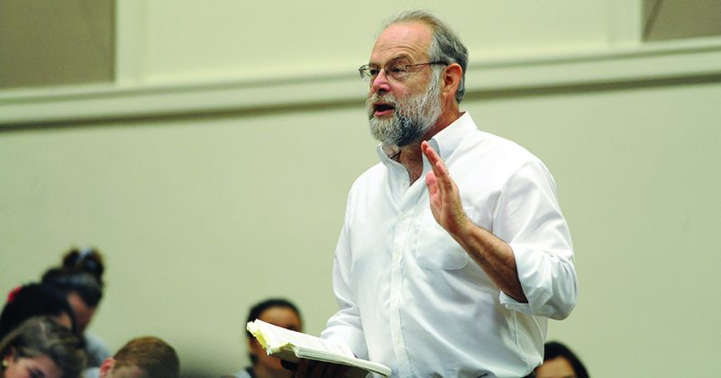 Geoffrey Sayre-McCord holding a book and gesturing while lecturing. Students sitting in the lecture hall visible in the background.