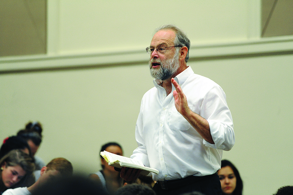Geoffrey Sayre-McCord holding a book and gesturing while lecturing. Students sitting in the lecture hall visible in the background.