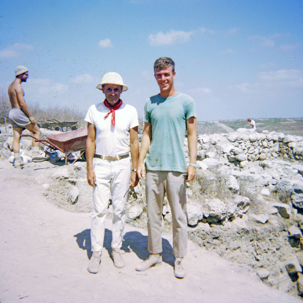 Bernard Boyd and Bill Farthing stand at an archaeological excavation site, with two other members of the dig and a red wheelbarrow visible in the background.