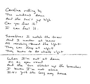 Handwritten lyrics to Carolina Rolling By song read: "Carolina Rolling By, the window's down and the sun's up high. Can you feel it? I can feel it. Sometimes I watch the trees, and I wonder at the leaves. All reaching toward the light. They can sleep at night. They know to do what's right. When I'm out at dawn on an open road, And the sun climbs up the branches, I know I'm not alone. It's just the long way home."