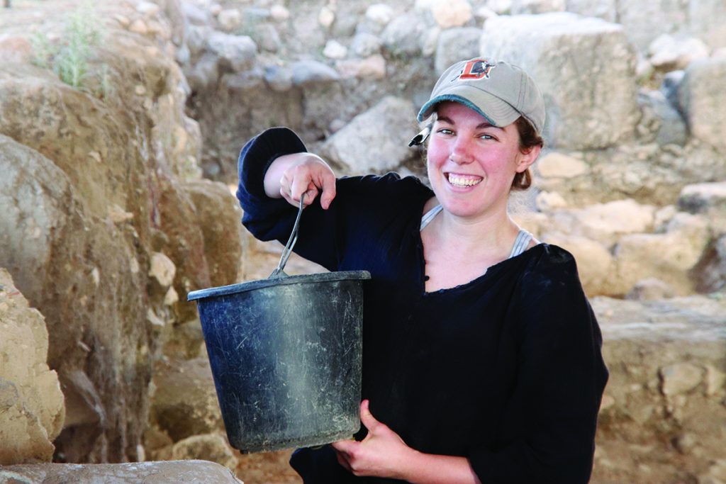 Jocelyn Burney holds a bucket of dirt as she smiles at the camera from the Huqoq dig site.