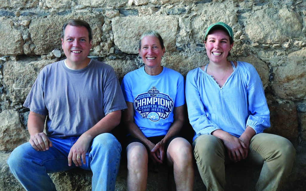 Sitting side by side against a stone wall and smiling at the camera are: Matthew Grey, Jodi Magness and Jocelyn Burney.