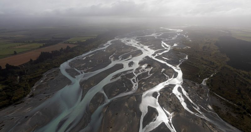 Overhead view of the sprawling Waimak river, trees and fields visible on either side and a hazy cloud layer at the top.