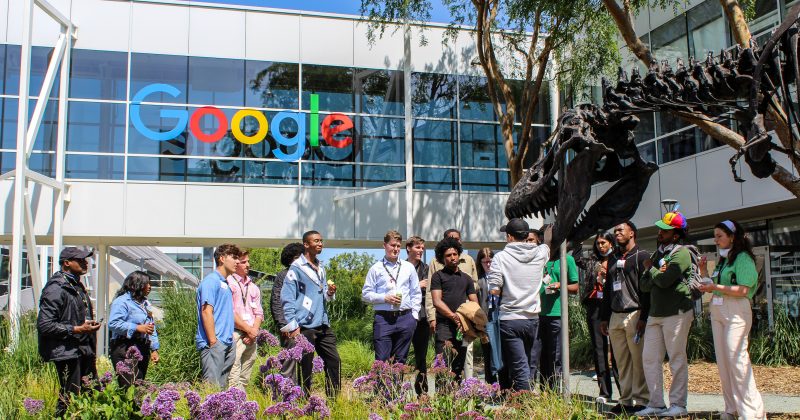 The Silicon Valley Maymester class stands under a "Google" sign on the Google campus. Their tour guide speaks about a large dinosaur skeleton – a T Rex – that is a focal point of the campus tour.