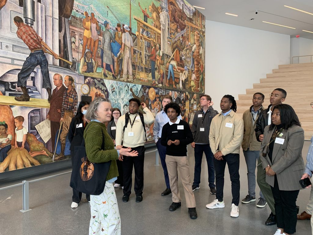 Students gather around a lecturer in front of a floor-to-ceiling fresco in the San Francisco Museum of Modern Art.