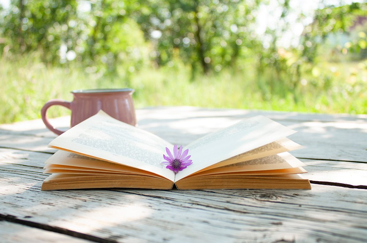 An open book with a small flower sitting in the center sits beside a white coffee cup on a table. Spring trees are in the background.