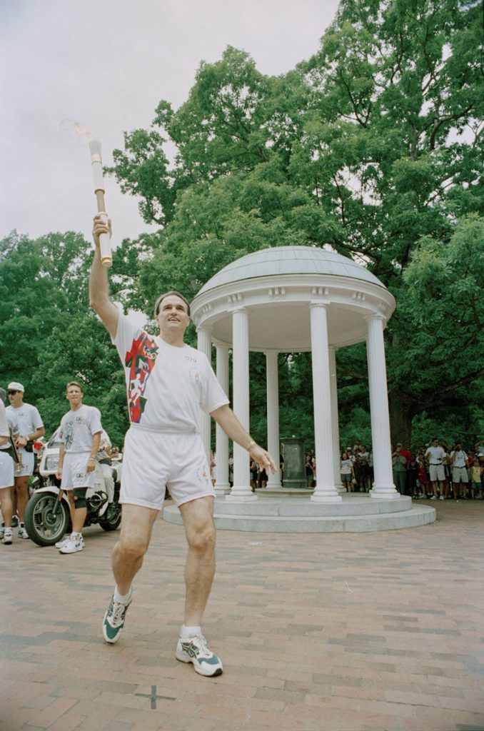 Charles Shaffer carries the Olympic torch in front of the Old Well, behind which a large crowd is gathered.